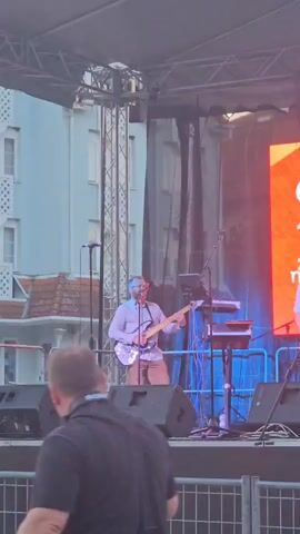 A man with glasses and a beard wearing a light blue shirt and brown pants plays a black bass guitar on a stage. He is standing in front of a microphone and several speakers are visible in the foreground. A large screen with an orange design is behind him. The background shows buildings and a metal structure.