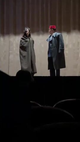 The scene depicts two actors on a stage. The actor on the left is cloaked in a full-length, dark olive green garment. The actor on the right wears a navy-blue suit with a white shirt and tie under a long, charcoal-grey coat. A red fez sits atop their head. The stage is in front of a wavy, brown-toned backdrop. An audience is visible in the dark foreground, with only the top of heads and some seats visible.