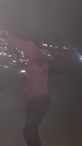 A blurry video shows a man in a maroon jacket and dark pants dancing in front of a city at night. The city lights are out of focus in the background. The man is moving his arms and legs, seemingly dancing to music that can be heard in the video. The scene is dark and the image is not very clear.