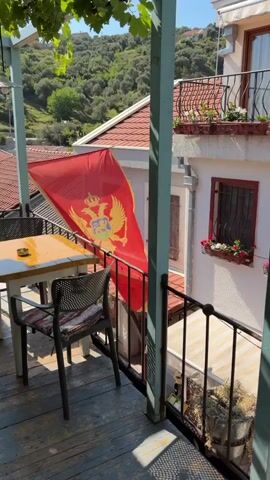 This video shows a view from a balcony overlooking a narrow street and buildings in what appears to be a coastal town. A Montenegrin flag is visible on the left side of the screen. A balcony with tables and chairs is featured prominently, with lush green vines growing over it. In the background, people are seen walking on a stone-paved street, and buildings with red-tiled roofs are visible. The overall atmosphere is relaxed and suggests a summer day.