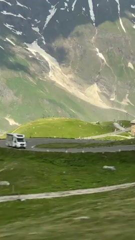 A white camper van drives along a winding mountain road. The road curves around a grassy slope, with a stone building situated on the right side of the road. Behind the road and building, a majestic mountain range with snow-capped peaks dominates the background. The mountains are a mix of rocky, barren terrain and lush green slopes. The sky is partly cloudy, with sunlight casting shadows on the mountains. There are other vehicles visible on the road, including what appears to be a tour bus parked near the building.