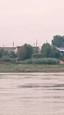 A panning video shows a wide river in the foreground and a shoreline with houses and trees in the background. The houses are small, some with colorful roofs, and appear to be part of a village. There are trees of various sizes scattered among the houses. Boats are visible on the river near the shore. The sky is overcast. The video is shot from a moving perspective, suggesting it was taken from a boat or vehicle.