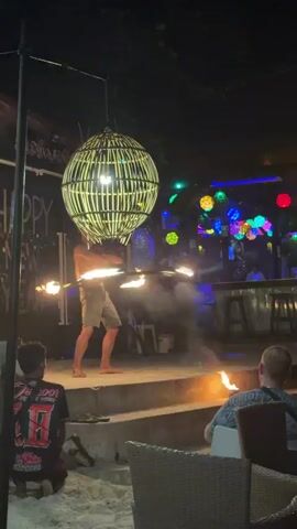 A medium shot captures a fire dancer on a stage at night, surrounded by people, lights, and objects. The male dancer, shirtless and wearing light-colored shorts, is positioned center-left on the stage, expertly manipulating a flaming hoop around his waist. He then shifts to hold a flaming stick in each hand, twirling them. To the left, a large woven spherical lantern hangs from above, partially obscuring the "HAPPY NEW YEAR" sign in the background. In the foreground, two individuals are seated at tables on a sandy ground, observing the performance. The person on the left is partially visible, while the person on the right is seen from behind, holding a phone. The background features a bar area adorned with numerous colorful spherical lights and glowing neon signs. A hazy smoke effect emanates from the fire dancer's act, adding to the atmosphere. The overall scene is lively and vibrant, with the fire performance serving as the central attraction.
