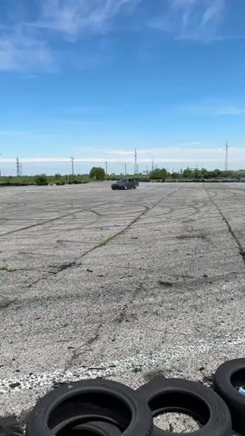 The video shows a dark gray sedan drifting on a large, gray asphalt surface. The surface is cracked and worn, with numerous dark tire marks indicating previous drifting activity. Several stacks of tires are visible in the foreground, near the camera. The car is the sole moving object, performing a wide, sweeping drift, kicking up a noticeable amount of white smoke from its tires. The background features a clear, blue sky with scattered white clouds. In the distance, there are utility poles and green vegetation lining the perimeter of the asphalt area. The video is shot from a stationary position, capturing the car's movements and the surrounding environment.