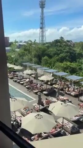 The video shows an aerial shot of a resort pool area, crowded with people sunbathing on lounge chairs under umbrellas and in cabanas. Some are swimming in the pool.  The area is bordered by trees and buildings.  A large communication tower rises in the background against a blue, partly cloudy sky. The view is from inside a building, looking out a window.