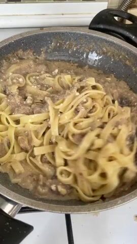 The video shows a close-up of a frying pan on a stovetop where fettuccine pasta is being mixed with a creamy sauce containing ground meat and possibly mushrooms. A wooden spatula is used to toss the pasta, coating it evenly with the sauce. The pasta is a bright yellow, and the sauce is a light brown color with visible pieces of meat and darker specks, likely pepper. The pan is dark gray and appears to be non-stick. The background shows a tiled backsplash and the white edge of a stovetop. The lighting is bright and appears to be natural. The camera is positioned slightly above the pan, looking down. The focus is sharp on the pasta and sauce as they are being stirred.