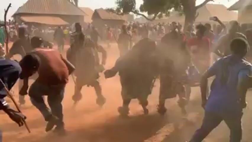 The video shows a crowded outdoor scene, possibly a village or community gathering, with several individuals dressed in elaborate, costume-like outfits, possibly representing traditional figures or characters. The individuals are kicking up dust as they move. Many people in the background appear to be onlookers, dressed in casual clothing. The ground appears to be dirt or reddish soil. Some people are carrying sticks. The overall atmosphere seems festive or ceremonial.