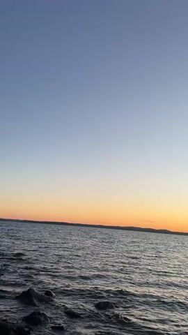 The video shows a wide shot of a body of water at sunset. The sky transitions from a clear, light blue at the top to a gradient of orange and yellow near the horizon. The water is dark and appears to have small waves. In the foreground, dark, jagged rocks are visible along the edge of the water. The horizon is flat and defined by a dark landmass in the distance. The overall atmosphere is calm and serene.