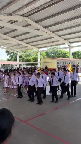 A group of young people in matching uniforms, boys in white shirts and black pants, and girls in light purple dresses, are performing a synchronized dance in an outdoor courtyard. The courtyard has a covered roof supported by white pillars. In the background, more people are watching the performance, and there are school buildings. The ground is paved with a dark surface marked with red lines. The overall atmosphere appears festive and organized, possibly a school event or performance.