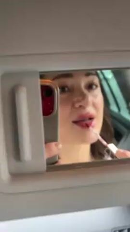 A woman applies lip gloss in her car using the sun visor mirror. She holds a phone to record herself. She smiles as she applies the lip gloss.