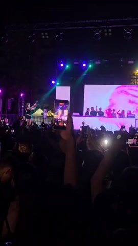 A crowd is gathered at an outdoor concert at night. The stage is lit with purple and green lights. A woman in a blue dress is singing and dancing on stage. A large screen behind her displays a pink and purple abstract image. Several people are standing behind a table on the stage, possibly part of the band or crew. In the foreground, several audience members are holding up their phones to record the performance. The audience is silhouetted against the stage lights, and their hands are raised in the air.