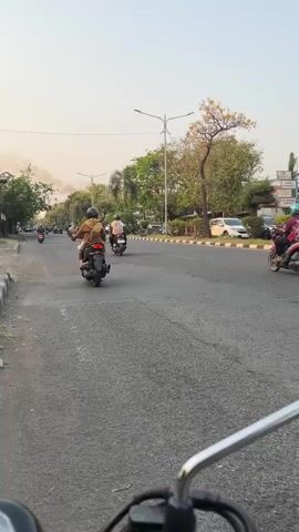 The video shows a view from a motorcycle on a paved road with several other motorcycles and a car. The sun is setting, casting a warm glow. On the left side of the road, there are buildings and trees. On the right side, there are more trees and a sidewalk with a white border. Several people are riding on the motorcycles, some with helmets, and some are wearing casual clothing. The car is black. There is a plume of smoke visible in the distance on the left side of the road. The road is in good condition with some cracks. The video is taken from a first-person perspective, suggesting the viewer is riding one of the motorcycles.