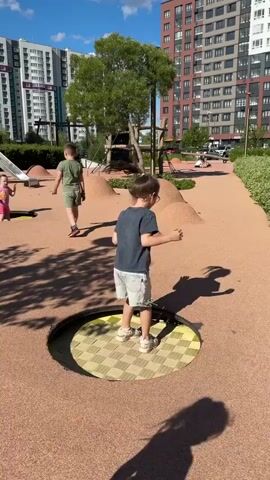 A young girl with brown hair in a ponytail, wearing a dark t-shirt and light-colored shorts, is shown playing on an outdoor playground. She is standing on a sunken circular trampoline with a checkered pattern. The playground has a sandy surface and hills made of sand. In the background, there are tall apartment buildings, trees, and other playground equipment, including a slide. Another child, a boy, is walking in the background. The sun is shining, casting shadows. The girl bounces on the trampoline and appears to be laughing.