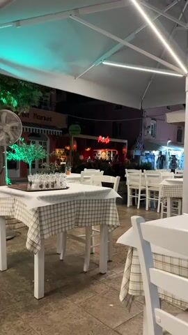 The video shows an outdoor restaurant or bar area at night. Several tables are set with white tablecloths and chairs. There is a DJ booth in the background with a person standing behind it. Neon signs are visible, including one that says "Pivoto". There are also plants and artwork in the background. The lighting is dim and colorful, with green and pink lights illuminating the scene.