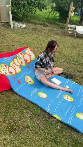 A young woman is sitting on a blue and red patterned mat outdoors, reading a book. She is wearing a black and white plaid shirt, tied at the waist, and light grey shorts. Her hair is wet, suggesting she may have recently swum or showered. She is holding the book open with her left hand and appears to be turning a page with her right hand. Next to her on the mat is a pack of cigarettes.

The setting is a grassy area, possibly a yard or a park, with trees and foliage in the background. A greenhouse or similar structure is visible to the left, and a wooden structure, possibly a fence or part of a shed, is to the right. The lighting suggests it is daytime, likely late afternoon or early evening. The overall mood appears relaxed and casual, with the woman engrossed in her reading. The visible text is on the cigarette pack, which appears to have Cyrillic lettering. The location is identified as Kuti, Russia.