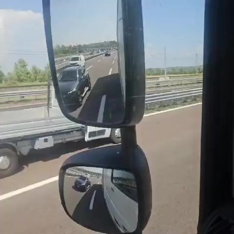 The video appears to be taken from the driver's perspective of a vehicle, likely a truck, on a highway. The camera is focused on the side mirrors and the road ahead.

In the main mirror, a black SUV is visible in the lane next to the camera vehicle. The SUV is in motion. Further down the highway, other vehicles, including a white van and several trucks, can be seen.

The smaller, lower mirror reflects the road behind the camera vehicle. A white van is visible in this reflection.

The environment is a sunny day on a multi-lane highway with a barrier on the left and trees and hills in the distance on the right. The road is paved and marked with white lines.

A GPS device is mounted on the dashboard in the foreground.

The video is shot in segments, with the view shifting slightly between the mirrors and the forward-facing view. The total duration of the video is approximately 5 seconds.

There are no people visible in the video, other than possibly the driver of the camera vehicle, whose presence is implied by the point of view.
