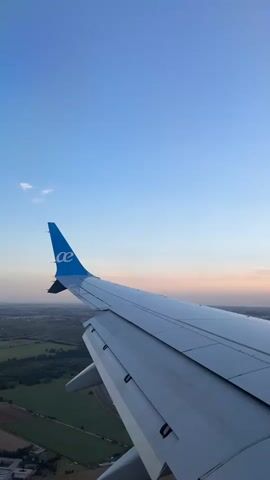 The video shows a view from inside an airplane, looking out the window at the wing and tail. The tail has a blue logo. The airplane is flying over land with trees, fields, buildings, and roads. There are cars on the roads. The sky is blue with some clouds, and the horizon has a pinkish hue. The scene is bright and appears to be during daytime.