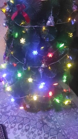 A small artificial Christmas tree is shown from an overhead perspective. The tree is decorated with colorful snowflake-shaped lights, bells, and a red bow. The lights are yellow, blue, green, and red. The background is a rug with a white and blue pattern. Christmas music is playing.