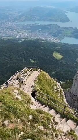 A drone shot begins by focusing on a mountain path with a wooden railing. A person wearing dark clothing walks down a set of stairs on the path. The drone then pans upwards and to the right, revealing a vast landscape. In the foreground, green grass covers a rocky mountainside. Beyond the mountain, a town or city is visible, nestled along the shores of a large, calm blue lake. Further in the distance, more bodies of water and rolling hills can be seen under a hazy sky. The scene appears to be a scenic vista from a high elevation.