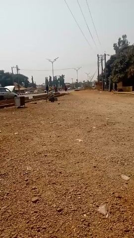 The video shows a wide shot of a dusty, reddish-brown gravel road or parking lot with some litter on the ground.  In the background, there is a paved road with some vehicles and pedestrians visible.  There are lampposts lining the paved road, and power lines overhead.  To the left, there are cars parked on the side of the road and a motorcycle parked on the gravel. A person is riding a motorcycle across the gravel area towards the paved road. There are trees in the background and a gate structure on the right side of the scene. The sky is hazy and bright.