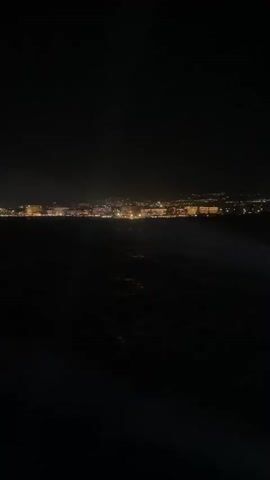 The video shows a dark body of water with lights of a city in the distance. There are some small illuminated objects or lights on the surface of the water in the foreground. The lights in the city appear to be from buildings and streetlights, creating a glowing horizontal line across the horizon. The overall impression is a nighttime scene with a distant urban landscape viewed from the water.