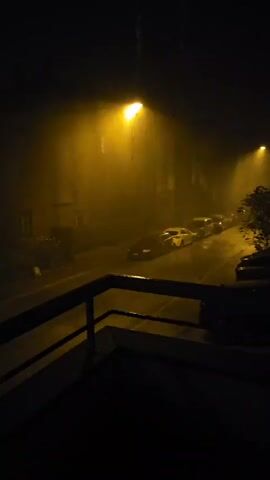 The video shows a dark, rainy street scene at night. A street lamp casts a yellow glow on the falling rain, creating a misty atmosphere. Several cars are parked along the side of the road, their reflections visible on the wet asphalt. A railing in the foreground suggests the footage is taken from a balcony or elevated viewpoint. The buildings lining the street are dimly lit. The rain appears to be falling steadily.