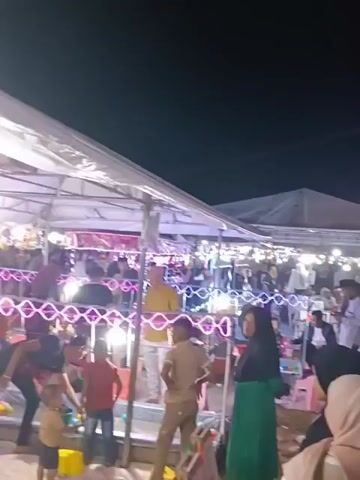The video captures a lively outdoor night market scene. A Ferris wheel, adorned with colorful lights, dominates the background. Below, a crowd of people navigates through stalls and rides, illuminated by strings of lights and the glow of various attractions. The atmosphere appears festive and bustling, with individuals and families enjoying the evening.