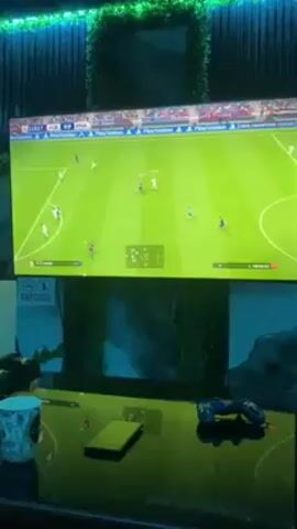 A video shows a soccer game on a TV screen. The game is between FCB and RMA, and the score is 0-0. The game clock reads 115:35. Players in white uniforms are on the offensive side near the FCB goal. A controller is on a black table, along with a green cell phone and a blue and white mug. There is also a picture on the table under the tv screen. A no alcohol or smoking sign is also on the table. The wall behind the television is decorated with green leafy plants.