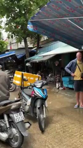 The video shows a street market scene with numerous vendors seated on the ground, selling fresh produce under blue and white striped awnings. Several people are riding motorbikes through the market, navigating between the stalls and vendors. The street is lined with green trees and the sky is overcast. The general atmosphere is busy and lively.