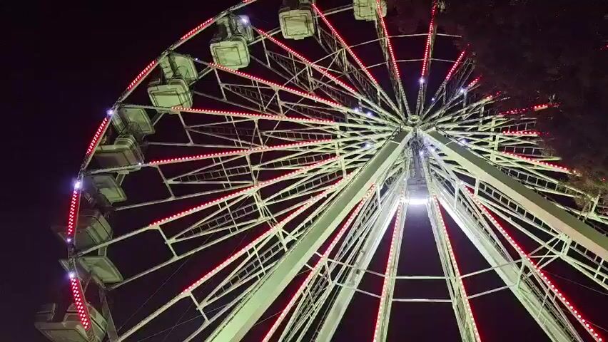 A Ferris wheel rotates against a dark night sky. The Ferris wheel is illuminated by changing colored lights that cycle through red, green, and blue. The structure of the Ferris wheel is visible, with its spokes and gondolas.