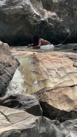 A person with dark, long hair, wearing a white cloth around their waist and no shirt, is crouching in shallow, flowing water next to a rocky waterfall. The person is reaching into the water with both hands. The environment is a natural setting with large, wet rocks and trees visible in the background.