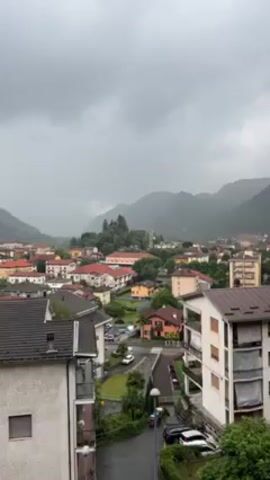 The video shows a cityscape with buildings of various sizes and colors, surrounded by mountains shrouded in mist. The sky is overcast and gray, suggesting a rainy or stormy day. There are cars parked on the streets, and the overall impression is one of a peaceful, yet slightly gloomy, urban setting.