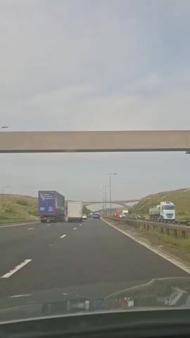 The video is a first-person view from a vehicle driving on a multi-lane highway. The sky is overcast. There are several large trucks and cars on the highway. A concrete overpass is visible in the foreground, and a bridge is seen further ahead. The road appears to be in a rural or semi-rural setting, with grassy hills on either side.
