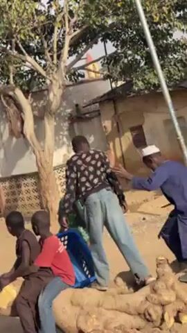 In a daytime outdoor setting, a group of children and men interact on and around a large tree root. Several children are seated or climbing on the root. One boy wears a red shirt and jeans, another a brown top and jeans. A blue plastic crate sits near them. A man in a black and white patterned sweater and blue jeans is standing on the root, being assisted by another man in a blue robe and white cap. A woman in a green top and headscarf is also nearby. The background includes a building with a corrugated metal roof and a tree with sparse foliage. The ground appears to be sandy and uneven. A light pole is visible. The camera moves to a wider view, revealing the surrounding area with more houses and a dirt road. Other people are seen in the distance. Some debris and construction materials are scattered on the ground.