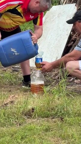 Two men are outdoors in a grassy area. One man on the left, wearing a red, yellow, and black shirt, is holding a large blue plastic container and pouring a brown liquid into a smaller blue-capped plastic bottle. This bottle is then placed into the opening of a larger, clear plastic bottle which is already partially filled with the same brown liquid. The man on the right, wearing a grey shirt and shorts, is assisting by holding the smaller bottle steady. In the background, there is a white object and some debris. The ground is covered in grass and dry leaves.