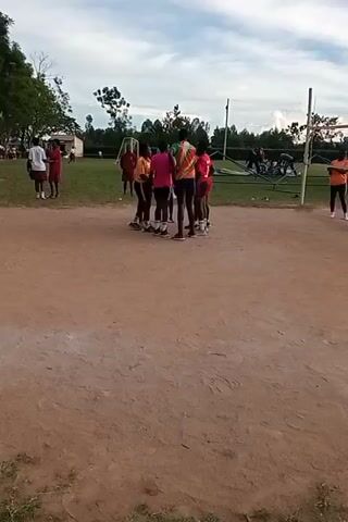 The video shows a group of young people standing in a circle on a dirt field next to a volleyball net. Some are wearing orange shirts, and one is wearing a pink shirt. A person in an orange shirt walks away from the group, stretching their leg. In the background, there are other people, a soccer net, trees, and a large tent. The sky is cloudy.