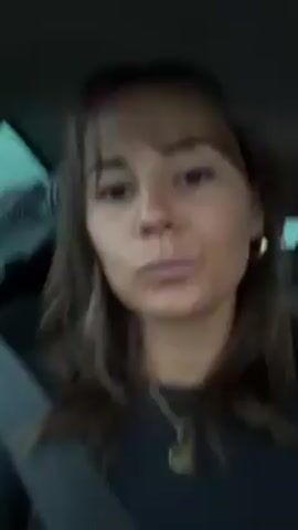 A young woman with brown hair and bangs is sitting in the driver's seat of a car. She is wearing a black top, a gold necklace, and gold hoop earrings. She is looking at the camera and making a face. A seatbelt is visible across her chest. In the backseat, a person is lying down and appears to be sleeping. The car is in motion.