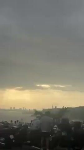 Here is a detailed description of the video:

The video shows a landscape view with a silhouetted cityscape on the horizon under an overcast sky. The sky is mostly gray with hints of yellow light breaking through the clouds. In the foreground, there are trees and foliage creating a dark outline against the brighter sky. People are present but obscured by the foreground elements. As the video progresses, the camera pans slightly, revealing more of the trees and the silhouette of the city in the distance. A white vehicle passes along the bottom of the frame toward the end of the video. The overall mood is atmospheric and somewhat muted due to the cloudy lighting.