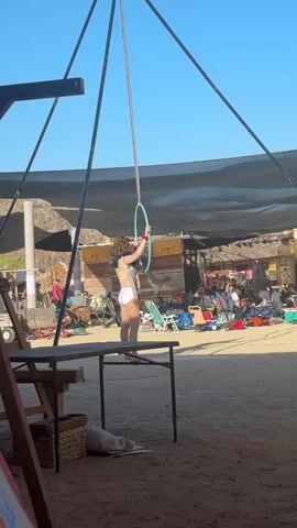 A medium shot, captured during daylight, shows a woman practicing with aerial hoops in an outdoor setting. The woman, dressed in a white leotard, stands on a table and is about to grab a hanging blue hoop. The hoop is suspended by a rope attached to a tent-like structure above. The backdrop reveals a bustling fair-like environment, filled with people, tents, chairs, and various objects. The lighting is bright, and the background is slightly blurred, focusing attention on the woman and the aerial hoop.