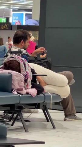 The video shows a couple sitting on airport benches. The woman is asleep with her head on the man's shoulder, wrapped in a blanket and scarf. The man is awake and looking at his phone. In the background, other travelers are seen sitting and walking around the airport waiting area. The environment appears to be a modern airport terminal with bright lighting and large windows. Another shot shows a person's legs and feet wearing sneakers, with the rest of the airport in the background.