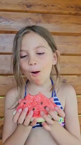 A young girl with light brown hair, wearing a blue and white striped bikini with butterfly patterns, is holding a slice of watermelon. She is looking to her left with a slight smile and her mouth slightly open, as if chewing or about to take a bite. The watermelon is red with black seeds. She is standing in front of a wooden wall, possibly outdoors given the lighting. The image appears to be taken during the daytime.