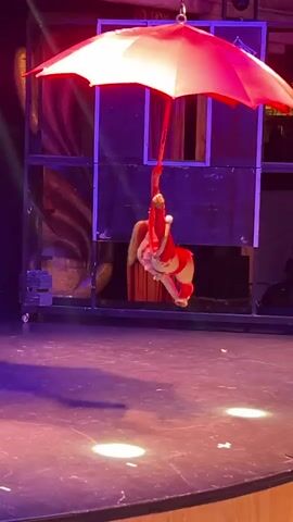 The video shows an aerial performer in a red costume suspended from a red and white umbrella. The performer is holding onto a rope attached to the umbrella's handle. The performer is in mid-air above a stage, with spotlights on the stage floor. In the background, there is a stage set with draped fabric and scaffolding. Two people are visible in the background, watching the performance. The lighting is dim and theatrical, with purple and red hues.