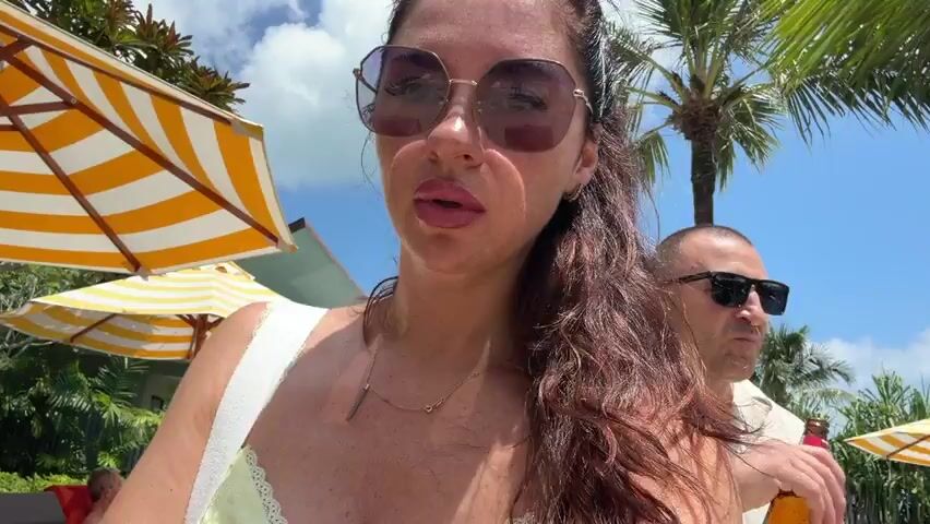 A medium shot focuses on a woman with dark hair, wearing sunglasses and a yellow dress, standing outdoors by a pool. To her right, a man in a patterned shirt walks past. In the background, there are palm trees, lounge chairs, umbrellas, and a swimming pool with people around it. The sky is blue with some clouds.