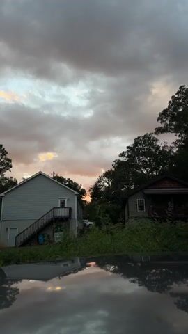 The video shows a view of two houses from a low angle, likely from the hood of a car. The sky is cloudy with hints of orange and pink from a sunset. The houses are surrounded by overgrown green grass and trees. The left house is light gray and has a set of stairs leading up to a door. The right house is dark green and appears to be older. The reflection of the sky and houses is visible on the surface in the foreground. There are no people or visible actions.