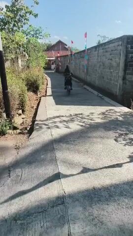 A short video clip shows a concrete road leading into a village on a sunny day. The road is lined with trees and a concrete wall. Several people are seen riding motorcycles on the road. Indonesian flags are displayed along the wall.