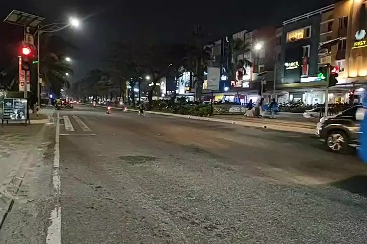 At nighttime, a blue bus is visible on the right side of the road. In front of the bus, two motorcycles are driving. The first motorcycle has a rider wearing an orange vest and a helmet. The second motorcycle has a rider wearing a dark jacket and a helmet. The road is paved and illuminated by streetlights and lights from buildings on the left side of the road. A traffic light is visible on the left side, showing red. There are palm trees lining the road.