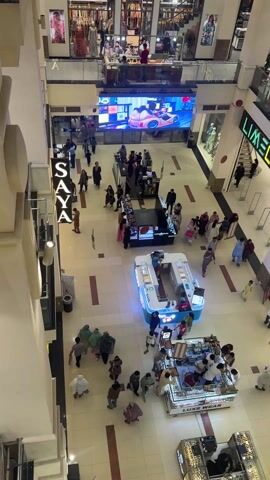 The video shows the interior of a multi-level shopping mall. People are walking around on the ground floor, and there are several small vendor kiosks in the center of the space.  A large LED screen shows graphics. Storefronts labeled "SAYA," "LIMELIGHT," and "DINERS" are visible. The video is taken from an elevated perspective.
