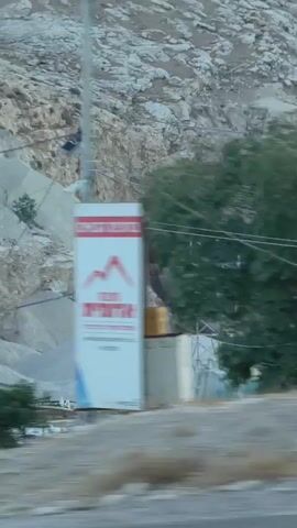 The video shows a side view of a moving vehicle traveling along a road. The road has a guardrail in the foreground. Beyond the guardrail, there is a dry, rocky, and sparsely vegetated landscape. Hills and mountains dominate the background, with some structures, possibly temporary housing or construction sites, visible on the slopes. The sky appears to be overcast or hazy, with soft lighting, suggesting either early morning or late afternoon. The motion blur indicates the camera is moving at a significant speed.