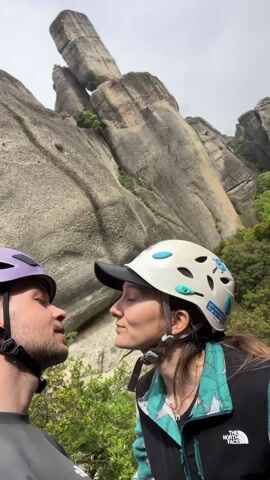 Two people, presumably a couple, wearing climbing helmets, are kissing in front of a large rock formation. The person on the left wears a purple helmet and a gray shirt. The person on the right wears a white helmet adorned with stickers and a jacket over a black top. The background consists of large, gray rock formations and some greenery. The sky appears overcast.
