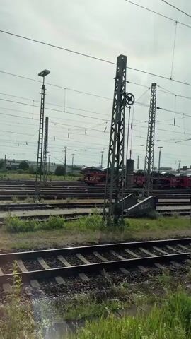 The video shows a railway setting featuring multiple parallel train tracks, overhead power lines, and numerous catenary poles. In the mid-ground, a freight train consisting of red or orange multi-deck car carrier wagons is slowly moving from right to left across the frame. The train is loaded with a variety of automobiles, including dark-colored sedans and SUVs, arranged on both upper and lower decks of the carriers.

The scene appears to be a rail yard or a railway siding. The sky is overcast, indicating a cloudy daytime. No people are visible in the video. There is no discernible text visible on the train or the cars it carries. The general impression is one of an industrial rail environment, likely near a car manufacturing or distribution hub, given the context of Ingolstadt, Germany.